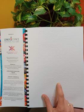 Pokaż zdjęcie: Osoba czyta miesięcznik „Help – jesteśmy razem” w wersji brajlowskiej. Palce leżą na stronie zapisanej alfabetem Braille’a. 