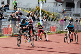 Pokaż zdjęcie: 4 osoby ścigają się na wózkach trzykołowych. Jedno koło jest z przodu dwa z tyłu. Nad siodełkami na wysokości klatki piersiowej są specjalne podpory. Wszyscy zawodnicy maja kaski na głowach.