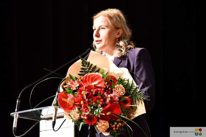 Pokaż zdjęcie: Przy mównicy stoi kobieta blondynka ubrana w ciemny garnitur. Na mównicy leży duży bukiet kwiatów.