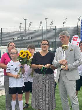 Pokaż zdjęcie: Kobieta i mężczyzna trzymają w rękach medal i puchar, obok stoi dziewczynka zawodniczka z kwiatami.