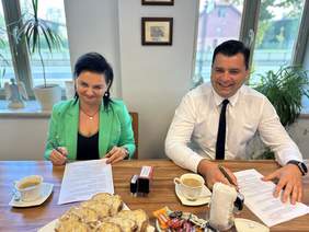 Pokaż zdjęcie: W jasnym pomieszczeniu, przy stole z poczęstunkiem, siedzą kobieta i mężczyzna. Oboje w wizytowych strojach. Pochylają się z uśmiechem nad dokumentami do podpisu. Za nimi widoczne są dwa, duże okna z widokiem na ulicę.