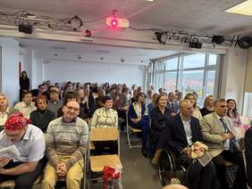 Pokaż zdjęcie: Sala pełna ludzi uczestniczących w wydarzeniu. W pierwszych rzędach osoby elegancko ubrane, jedna z kwiatami. Widoczne uśmiechy i zainteresowanie. W tle duże okna z widokiem na miasto. Atmosfera uroczysta i integracyjna.