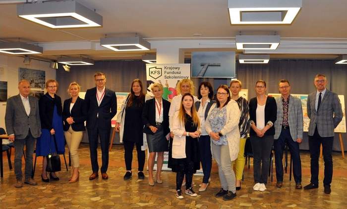 Pokaż zdjęcie: W dużej sali na tle wystawy prac plastycznych oraz banera Krajowego Funduszu Szkoleniowego stoi dziesięć kobiet i czterech mężczyzn, którzy pozują do zdjęcia.