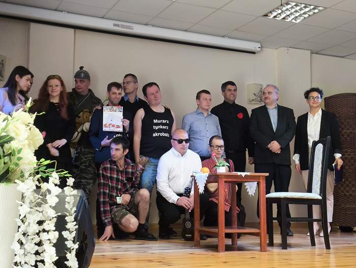 Pokaż zdjęcie: Grupowe zdjęcie uczestników wydarzenia teatralnego na scenie. Kilkanaście osób stoi i siedzi obok siebie, przed nimi mały stolik z dekoracją i krzesło. W tle jasna ściana i opuszczony ekran projekcyjny.