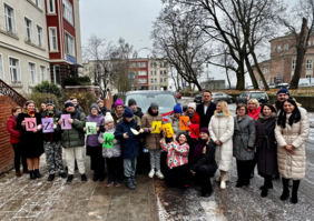 Pokaż zdjęcie: Duża grupa osób, dorosłych i młodzieży, stoi na chodniku przed ciemnym vanem marki Ford. Osoby trzymają duże, kolorowe litery, które razem układają się w słowo "DZIĘKUJEMY". W tle widać budynki mieszkalne, drzewa bez liści i ulicę. Na ziemi leży lekka warstwa śniegu. Grupa wydaje się pozować do zdjęcia w zimowy, pochmurny dzień.