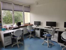 Pokaż zdjęcie: Pomieszczenie pracowni komputerowej z dużym oknem pod oknem stoi biurko z krzesłem obrotowym na nim stoi czajnik i urządzenie do laminowania pod ścianą przy oknie stoją trzy biurka na nich ustawione są monitory i klawiatura z myszką