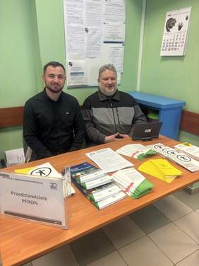 Pokaż zdjęcie: Dwóch uśmiechniętych mężczyzn znajduje się w pomieszczeniu z zielonymi ścianami. Przed nimi stół z licznymi ulotkami i broszurami. Po lewej stronie stojak z kartką z napisem: ,,Przedstawiciele PFRON’’.