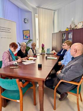 Pokaż zdjęcie: Zdjęcie stołu konferencyjnego przy którym po obydwu stronach siedzi łącznie sześć osób.