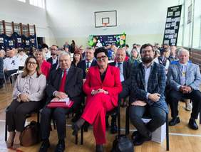 Pokaż zdjęcie: Grupa uczestników przeglądu poezji i piosenki siedzi na krzesłach w sali gimnastycznej. W pierwszym rzędzie znajdują się zaproszeni goście Monika Wac, Bożena Borowiec i Kamil Bobek. W tle widoczna jest dekoracja z kolorowymi kwiatami i napisem „Każdy z nas jest wyjątkowy”.