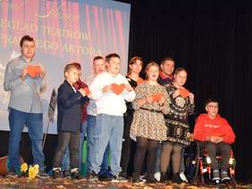 Scena z grupą osób stojących w rzędzie, trzymających czerwone, sercowate wycinanki. W grupie są zarówno dorośli, jak i dzieci, niektóre osoby mają widoczną niepełnosprawność, w tym chłopak w czerwonej bluzie siedzący na wózku inwalidzkim po prawej stronie. Za nimi duży ekran z napisem "Festiwal Słońca. II Przegląd Teatrów Niepełnosprawnego Aktora".