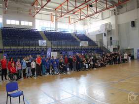 Pokaż zdjęcie: na zdjęciu hala sportowa, na sali duża grupa osób prezentuje swoje drużyny. Po środku siedząca na wózku pełnomocnik FAR przemawia do uczestników Spartakiady  