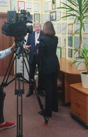 Pokaż zdjęcie: pomieszczenie sekretariatu w Oddziale w tle na ścianie wiszą dyplomy i podziękowania po prawej stronie stoi dyrektor i udziela wywiadu, po lewej jest umieszczona kamera 