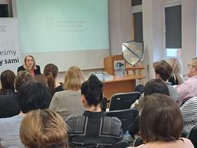 Pokaż zdjęcie: Kobieta siedząca za biurkiem, prowadzi prezentację w sali konferencyjnej, z logotypem PFRON na ekranie w tle w tle. Przed nią siedzą uczestnicy. Obok stoi sztaluga z tarczą z herbem.