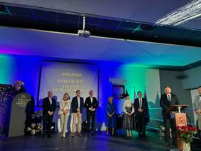 Pokaż zdjęcie: Na scenie trwają uroczystości z okazji obchodów jubileuszy. Przy mównicy stoi mężczyzna, który przemawia do mikrofonu. W tle stoją także inne osoby. Na ekranie wyświetlony jest napis: 30-lecie działalności Warsztatu Terapii Zajęciowej oraz 25-lecie Stowarzyszenia Na Rzecz Osób Niepełnosprawnych „RAZEM". 