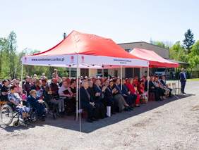 Uczestnicy spotkania grupa ponad 20 osób siedzą na ławeczkach pod dużym namiotem, słoneczny dzień.