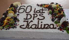 Pokaż zdjęcie: Duży prostokątny tort z napisem „60 lat DPS Kalina”, udekorowany owocami, kawałkami czekolady i bitą śmietaną, znajdujący się na białym obrusie.