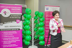 Pokaż zdjęcie: Na zdjęciu Krystyna Kronenberger - prezes Stowarzyszenia. Stoi na tle różowego roll-upu informacyjnego.