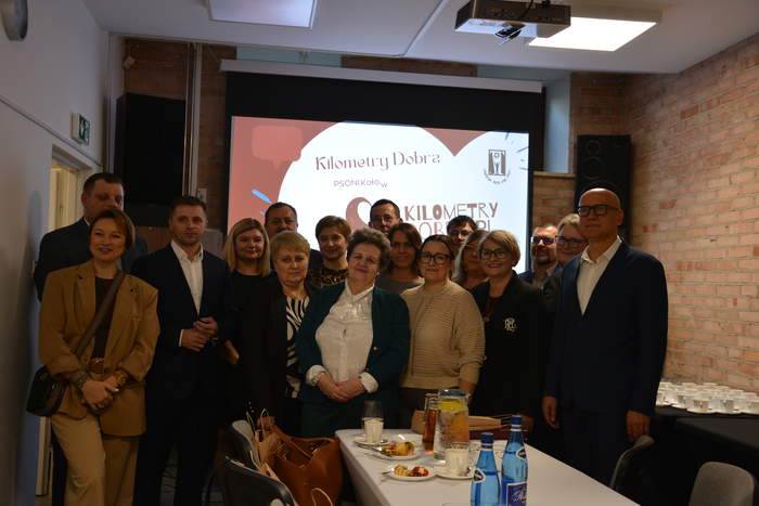 Pokaż zdjęcie: W jednej z sal na tle ekranu z prezentacją grupa osób pozuje do pamiątkowej fotografii.