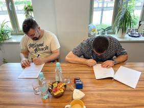 Pokaż zdjęcie: Przy stole siedzi dwóch mężczyzn ubranych w koszulkach z krótkim rękawem, podpisują dokumenty, na biurku leżą pieczątki.