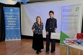 Pokaż zdjęcie: Dwie kobiety stoją na przodzie sali gimnastycznej, w tle banery z logo PFRON oraz Inspekcji Pracy.