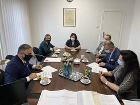 Pokaż zdjęcie: Przy ciemnym drewnianym stole siedzi trzech mężczyzn i trzy kobiety. Wszyscy mają założone maseczki, mężczyźni w marynarkach i koszulach kobiety w eleganckich sukienkach. Na stole leża podpisywane dokumenty, pieczątki a także woda mineralna, ka