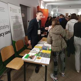 Pokaż zdjęcie: Grupa studentów na korytarzu uczelni, gdzie swoje stoiska mają wystawcy Targów Pracy. Z przodu stoisko Oddziału Wielkopolskiego PFRON, przy którym mężczyzna udziela informacji uczestnikowi Targów. Na stoliku wyłożone są materiały informacyjne.