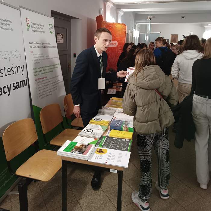 Pokaż zdjęcie: Grupa studentów na korytarzu uczelni, gdzie swoje stoiska mają wystawcy Targów Pracy. Z przodu stoisko Oddziału Wielkopolskiego PFRON, przy którym mężczyzna udziela informacji uczestnikowi Targów. Na stoliku wyłożone są materiały informacyjne.