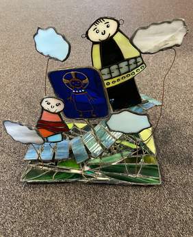 Pokaż zdjęcie: Trójwymiarowy witraż. Z szklanego, w odcieniach zieleni podłoża wystają szklane figurki uśmiechniętego księdza, pielgrzyma i obrazu świętego. Ponad nimi wystają na drucikach szklane chmurki.