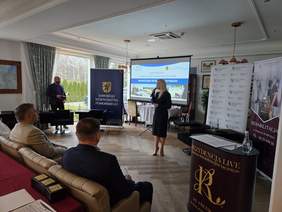 Pokaż zdjęcie: Sala konferencyjna. Kobieta przemawia przy mikrofonie, w tle ekran z prezentacją. Uczestnicy siedzą w eleganckich fotelach. Widoczne banery, m.in. Urzędu Marszałkowskiego Województwa Pomorskiego. Za oknem ogród. Atmosfera oficjalna.