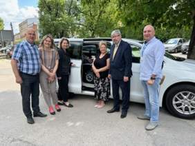 Pokaż zdjęcie: Przed samochodem stoi sześć osób: trzy kobiety i trzech mężczyzn. We wnętrzu auta znajduje się osoba siedząca na wózku inwalidzkim. 