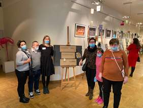 Pokaż zdjęcie: Fragment pomieszczenia, na wprost jest czarny karton z napisem wystawa uczestników WTZ Racibórz, z jednej i drugiej strony kartonu stoją trzy osoby w tle widać powieszone kolorowe obrazy i kobietę która je ogląda.