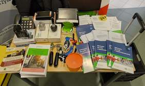 Pokaż zdjęcie: Widok na stół z ulotkami i sprzętem. Od lewej telefon dla osób słabowidzących, luba powiększająca i aparaty słuchowe, po prawej ulotki CIDON, OWiT i informator ZUS