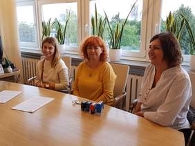 Pokaż zdjęcie: Trzy panie siedzą przy jasnym dużym stole nad dokumentami, za nimi ściana okien