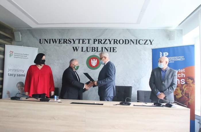 Pokaż zdjęcie: Cztery osoby stoją na tle ściany z napisem Uniwersytet Przyrodniczy w Lublinie. Dwie środkowe osoby wymieniają się uściskiem dłoni po podpisaniu wspólnego Porozumienia.