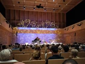 Pokaż zdjęcie: Scena w dużej sali koncertowej, Na senie widać instrumenty muzyczne. Na widowni goście.