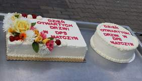 Pokaż zdjęcie: Zdjęcie dwóch tortów jeden z nich jest prostokątny udekorowany kwiatami, drugi okrągły bez kwiatów. Obydwa mają napis Dzień Otwartych Drzwi DPS Matczyn