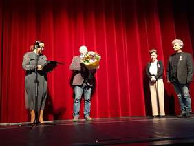 Pokaż zdjęcie: Scena z czerwoną kotarą na której stoją cztery osoby, przy mikrofonie kobieta, odczytuje list.