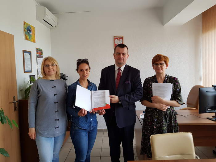 Pokaż zdjęcie: Sygnatariusze umowy stoją w pomieszczeniu, gdzie podpisywano umowę, na tle ściany w kolorze białym, widać m.in. godło Polski