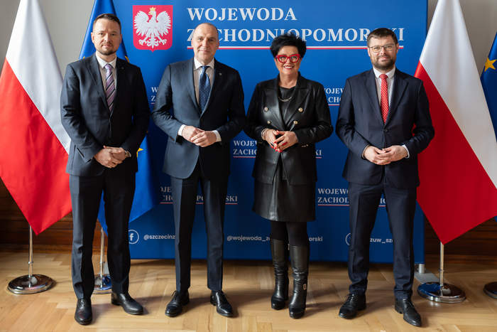 Pokaż zdjęcie: Na zdjęciu znajdują się cztery osoby – trzech mężczyzn i jedna kobieta – pozujące na tle niebieskiego banera z napisem „WOJEWODA ZACHODNIOPOMORSKI” oraz polskim godłem. W tle znajdują się także flagi Polski oraz Unii Europejskiej. Wszyscy są ubrani w formalne stroje – mężczyźni mają na sobie garnitury, a kobieta czarną skórzaną marynarkę i spódnicę, do których dobrała wysokie czarne buty. Kobieta wyróżnia się czerwonymi okularami i lakierem do paznokci w tym samym kolorze. Osoby stoją w lekkim rozkroku, z dłońmi splecionymi lub ułożonymi w naturalny sposób. Wszyscy patrzą bezpośrednio w obiektyw i mają neutralne lub lekkie uśmiechy.