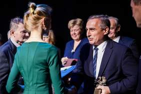 Pokaż zdjęcie: Mężczyzna w granatowym garniturze i białej koszuli krawacie w odcieniach niebieskiego trzyma statuetkę gryfa pomorskiego we jednym ręku, prawą dłonią ściska dłoń kobiety w sukience i blond spiętych włosach.