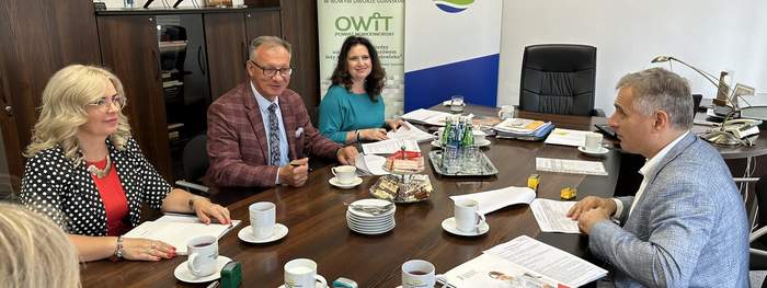 Pokaż zdjęcie: Przy stole w biurze siedzą dwaj mężczyźni i dwie kobiety, patrzą na siebie z uśmiechami. Na stole leż dokumenty pieczątki kawa. W tle baner Powiat Nowodworski i OWIT.