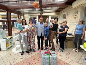 Pokaż zdjęcie: W rządzie stoją kobiety i mężczyźni, trzymają dzieci na rękach. Przed nimi wielki prezent leż na podłodze obsypanej konfetti.