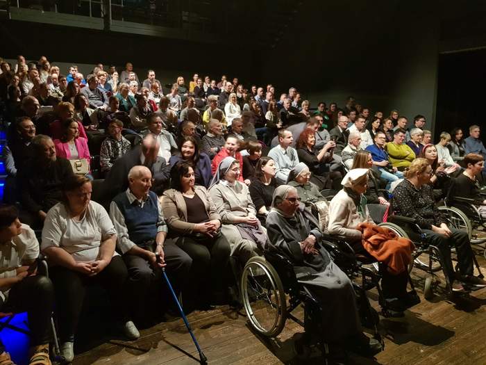 Pokaż zdjęcie: Grupa około 150 kobiet i mężczyzn, z czego część z tych osób porusza się na wózku, siedzi w sali teatralnej i oczekuje wyników III edycji Konkursu Oddziału Wielkopolskiego PFRON na wykonanie kartki bożonarodzeniowej przez osoby z niepełnosprawnością.