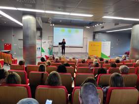 Pokaż zdjęcie: Zdjęcie sali konferencyjnej, zrobione z tyłu, widać rzędy czerwonych foteli, scenę i prelegenta.