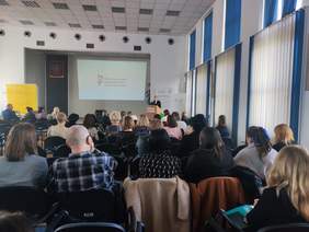 Pokaż zdjęcie: Zdjęcie z tyłu sali konferencyjnej w kierunku sceny, na przodzie przy mównicy stoi kobieta w ciemnym garniturze.