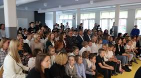 Pokaż zdjęcie: W dużej sali znajduje się grupa kilkudziesięciu osób, wśród których w przewadze są dzieci. Osoby te w większości siedzą, jednak część z nich przyjęła pozycję stojącą.