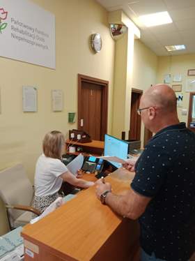 W sekretariacie Łódzkiego Oddziału PFRON znajdują się dwie osoby: kobieta i mężczyzna. Pani siedzi na krześle i patrzy w ekran monitora. Pan stoi przy ladzie i podaje jakieś dokumenty. Na biurku stoją także różne urządzenia biurowe. 