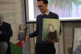 Pokaż zdjęcie: Mężczyzna w okularach stoi  i  trzyma w rękach prace plastyczne z fundacji IKAR, prezentując je uczestnikom konferencji
