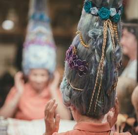 Pokaż zdjęcie: Na pierwszym planie głowa kobiety od tyłu z założoną bardzo dużą fantazyjną peruką z doczepionymi kwiatami, sznurkami i perłami. W tle jest jej niewyraźne odbicie w lustrze.