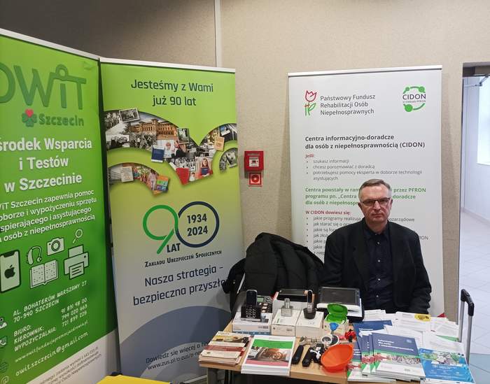 Pokaż zdjęcie: Z lewej strony widoczny rollup Ośrodka Wsparcia  i Testów z danymi adresowymi w kolorze zilonym, obok zielony rollup ZUS z informacją o 90-leciu. Na wprost biało zilony baner PFRON i CIDON, po środku siedzi pracownik w okularach, a przed nim stół na która znajdują się ulotki OWIT, PFRON i ZUS, a także sprzęt wspomagający - min. telefon i sztućce.  